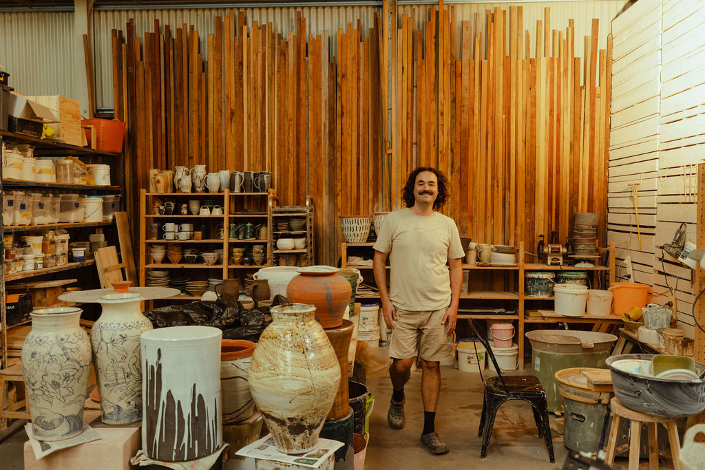 A portriat photography of Remy Pajaczkowski-Russell in his studio surrounded by ceramics and other pottery equipment